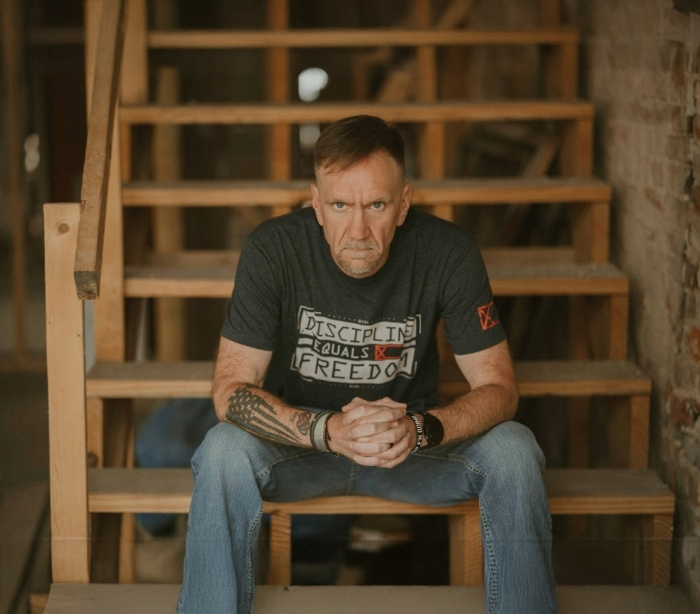 Jim Lunsford sitting on wooden steps in a focused pose, wearing a “Discipline Equals Freedom” shirt, representing strength, discipline, and recovery.