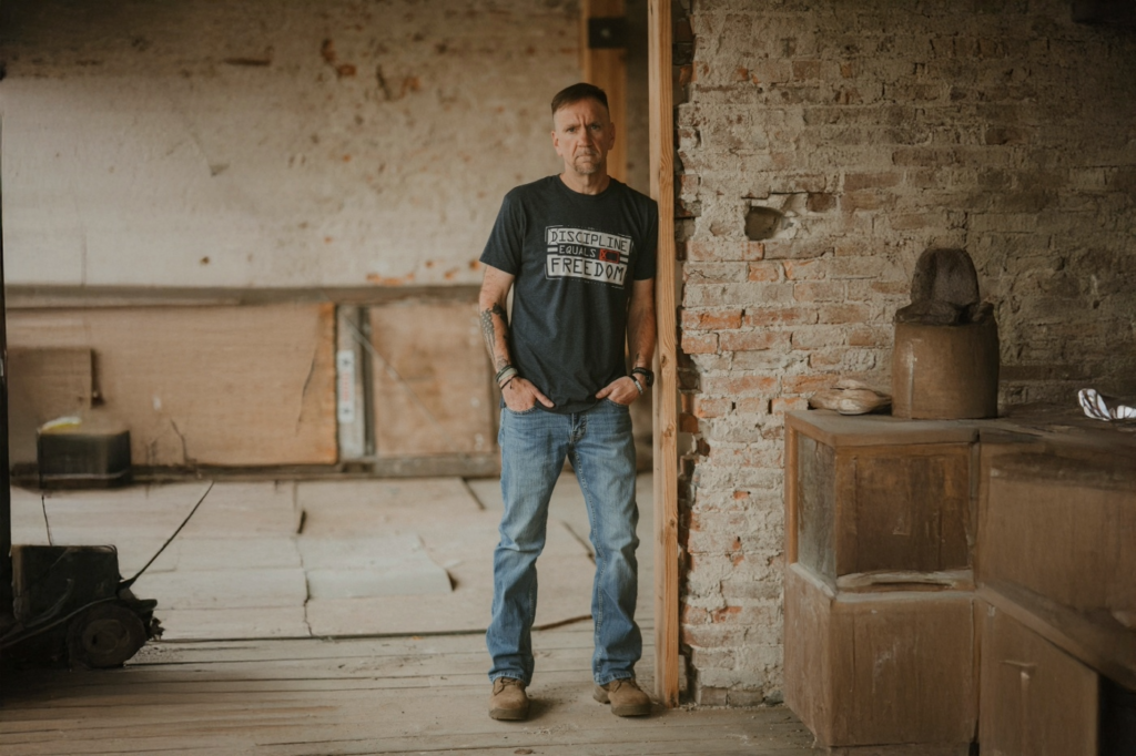 Jim Lunsford standing in a rustic room wearing a black ‘Discipline Equals Freedom’ shirt, symbolizing strength, recovery, and purpose.